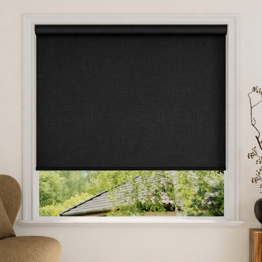 Black roller blind covering a window with a view of greenery outside.